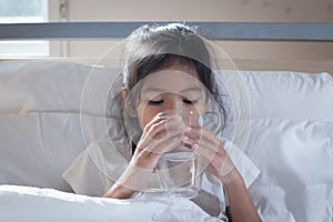 Sick asian child girl is drinking water from a glass after eat medicine in bedroom.