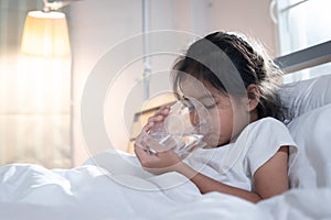 Sick asian child girl is drinking water from a glass after eat medicine in bedroom.