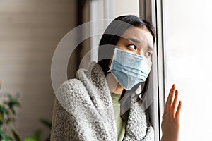 Sick asian girl in medical face mask standing by the window and yearning to go outside, being on quarantine, ill with