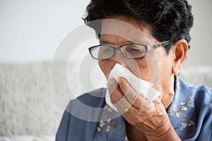 Sick asian old woman using tissue paper close mouth while cough, sitting on sofa at home. Senior healthcare concept