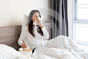 Sick Asian woman feeling cold and has a high fever blowing nose while sitting on bed at home. Health problems