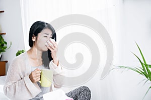 Sick Asian woman sneezing because of flu hand holding tissue and glass of hot water sitting in bed