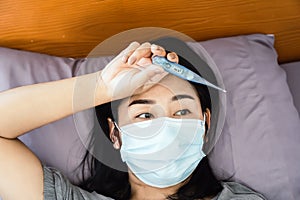 sick Asian woman wearing face mask hand holding thermometer having fever  high body temperature laying down in bed