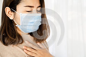 Sick Asian woman wearing face mask having sore throat from cold and flu standing next to the windows