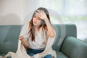 Sick Asian young woman sit under blanket on sofa she sneeze checking temp with digital thermometer