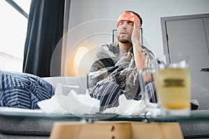 Sick bearded man who has bad cold or seasonal flu sitting on couch at home. Guy with fever wearing warm plaid shivering