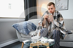 Sick bearded man who has bad cold or seasonal flu sitting on couch at home. Guy with fever wearing warm plaid shivering