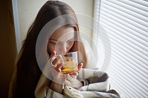 Sick beautiful woman with colds and drinks hot tea with lemon sitting by the window in a country house in winter