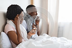 Sick black couple sitting in bed, measuring fever, sneezing noses