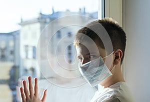 A sick boy in a medical mask stands at the window and looks longingly. photo