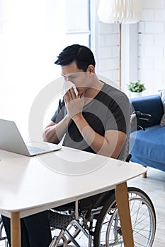 Sick Business entrepreneur in wheelchair sneezing in the home office. Disabled Sick Young Man on Wheelchair at Home. Young man in