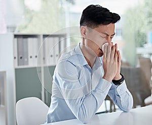 A sick businessman using a tissue and blowing his nose while working in a modern office. An ill asian man ill with flu