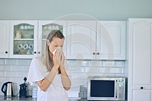 Sick caucasian woman holding tissue and blowing her nose while standing in kitchen at home. Woman suffering from