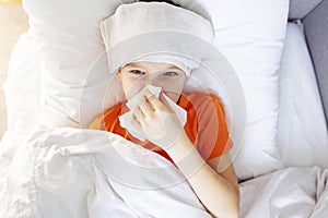 Sick child boy is lying in bed with high fever and blowing his nose, resting at home