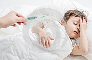 Sick child with flu fever laying in bed and mother holding thermometer