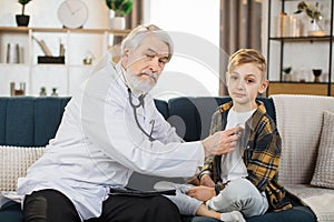 Sick child sitting on the couch, while caring confident mature male doctor listening his heartbeat