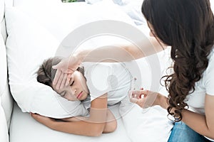 Sick cute teen girl lying in bed with fever while her mother sits near and checks the temperature holding termometer in