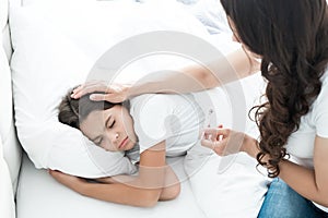 Sick cute teen girl lying in bed while her mother sits near and checks the temperature holding termometer in her hand