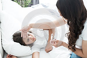 Sick cute teen girl lying in bed while her mother sits near and checks the temperature using termometer