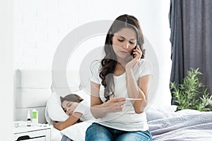 Sick cute teen girl sleeping in bed while her mother sits near and calls the doctor holding termometer in her hand photo