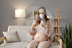 Sick displeased pregnant woman wearing medical mask sitting on sofa with medicine and smartphone using mobile phone searching