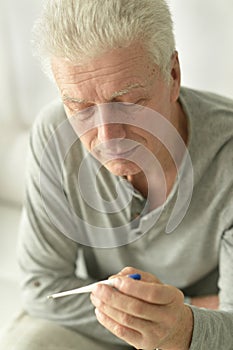 A sick elderly man sits on his bed and checks his body temperature.