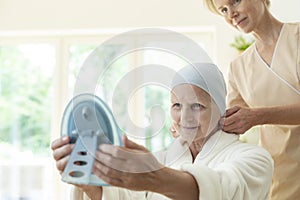 Sick elderly woman with headscarf and caregiver looking in the mirror