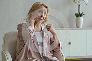 A sick elderly woman sneezes using a paper napkin for her nose, she has a runny nose and coughs sitting on a chair in