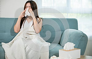 Sick female sitting under blanket on sofa and sneeze with tissue paper in living