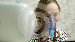 A sick girl in a mask inhales a spray bottle and plays a computer game on a tablet. Kid in a blue hospital mask inhale