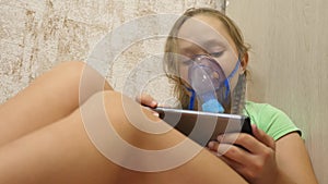 A sick girl in a mask inhales a spray bottle and plays a computer game on a tablet. Kid in a blue hospital mask inhale