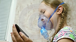 A sick girl in a mask inhales a spray bottle and plays a computer game on a tablet. Kid in a blue hospital mask inhale