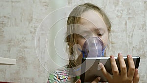 A sick girl in a mask inhales a spray bottle and plays a computer game on a tablet. Kid in a blue hospital mask inhale