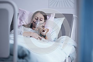 Sick girl with oxygen mask sleeping in a hospital bed with teddy