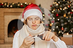 Sick girl in santa hat, wrapped in scarf, pouring cough syrup into spoon, looking at medicines and measuring temperature with