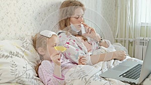 Sick girl with a temperature. Child with fever is lying in bed with her mother, eating fruit and using a laptop.