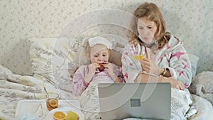 Sick girl with a temperature. Child with fever is lying in bed with her mother, eating fruit and using a laptop.