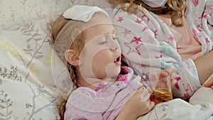 Sick girl with a temperature. Child with fever is lying in bed with her mother, eating fruit and using a laptop.