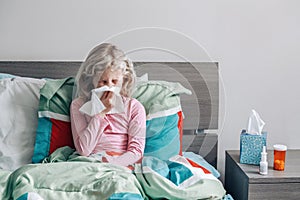 Sick ill Caucasian girl with fever lying in bed at home blowing nose with a napkin paper tissue. Virus cold season flu illness.