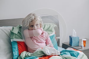 Sick ill kid girl with fever lying in bed at home with flu, fever, running nose. Child coughing sneezing in elbow. Virus cold