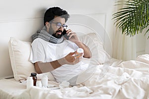 Sick Indian Man Holding Pills And Calling Doctor While Sitting In Bed