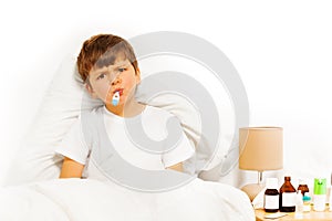 Sick kid boy sitting in bed and taking temperature