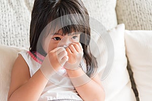 Sick little Asian girl wiping or cleaning nose with tissue sitting on sofa at home. Medicine and health care concept.