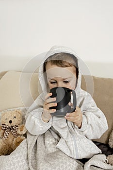 Sick little boy drinking hot tea on the bed at home. Unwell, illness child wrapped in a blanket, with mug in his room