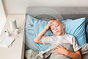 Sick little boy with high fever and headache laying in bed checking his forehead. Stay at home during corona virus epidemic