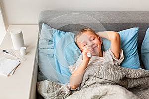 Sick little boy with high fever and headache laying in bed spraying medicine into his nose. Stay at home during corona virus