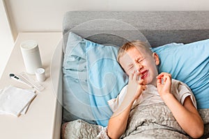 Sick little boy with high fever and headache laying in bed spraying medicine into his nose. Stay at home during corona virus