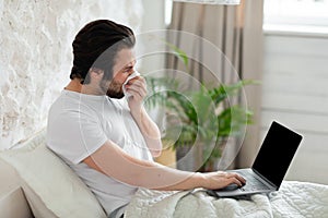 Sick man laying in bed, sneezing and using laptop, mockup