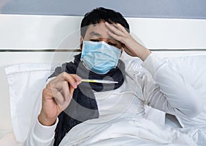 Sick man in medical mask headache and using thermometer to checking his temperature on bed, coronavirus covid-19 pandemic