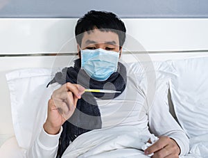 Sick man in medical mask using thermometer to checking his temperature on bed, coronavirus covid-19 pandemic concept